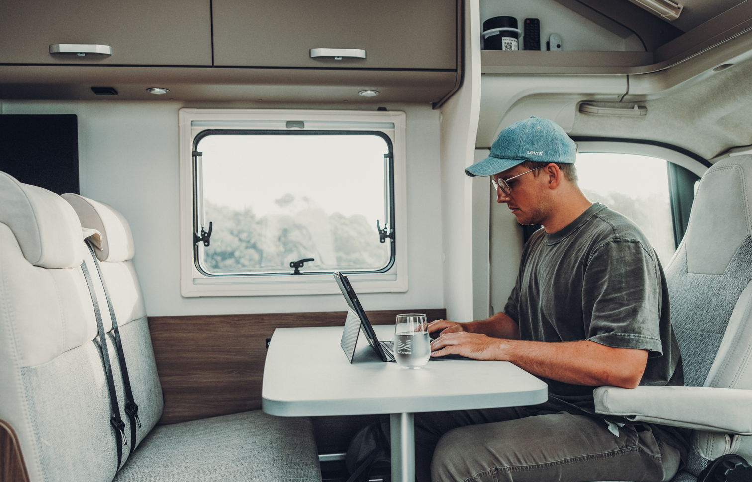 Homme travaillant sur un ordinateur portable dans un camping-car à une table, avec une banquette et une fenêtre en arrière-plan.