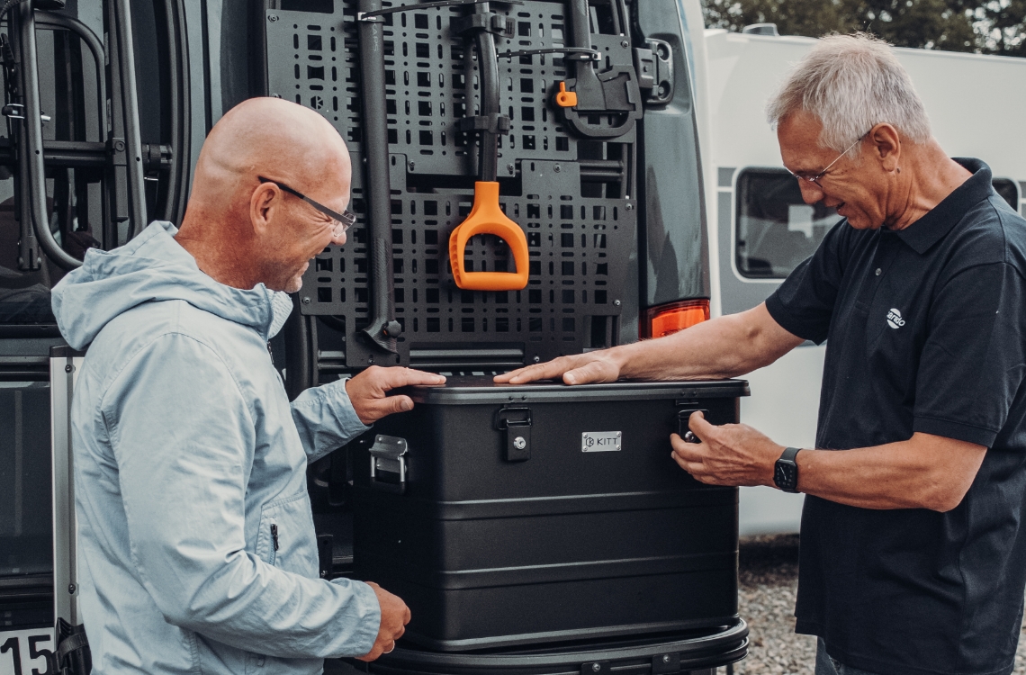 Dos personas fijan una caja de transporte al sistema de transporte trasero de una autocaravana.